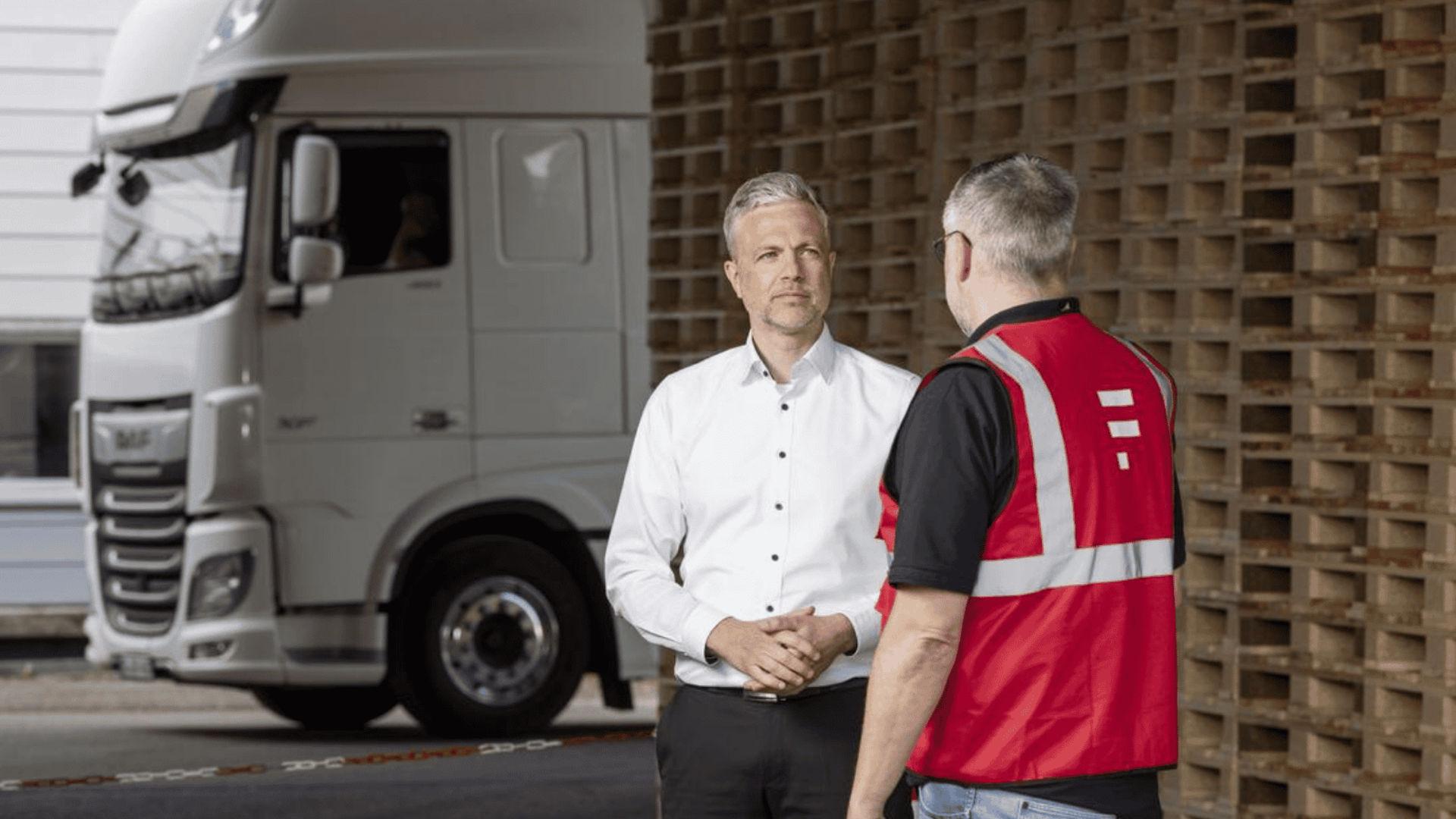 Zwei Personen im Gespräch vor einem LKW und einer Wand aus Paletten – Abstimmung in der Supply Chain